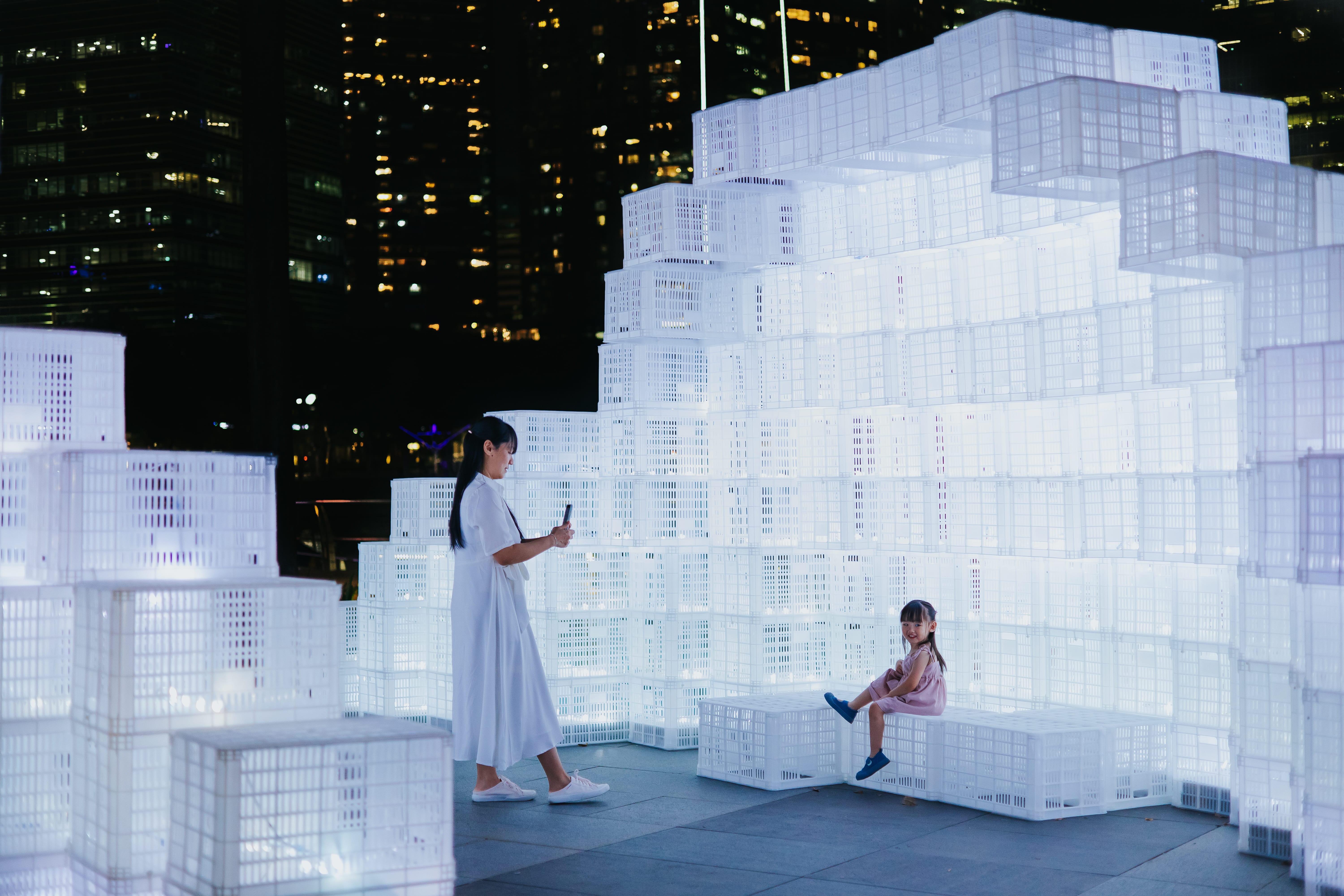 An arch-shaped structure built from stacked glowing white crates, with visitors exploring and sitting inside it in a dimly lit space.